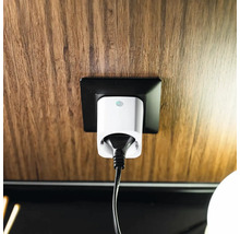 Weißer Smart-Home-Zwischenstecker mit schwarzem Kabel in einer schwarzen Steckdose an einer Wand mit Holzmaserung.