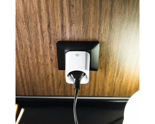 Weißer Smart-Home-Zwischenstecker mit schwarzem Kabel in einer schwarzen Steckdose an einer Wand mit Holzmaserung.