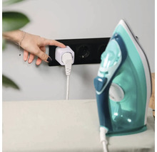 Eine Hand drückt die Taste an einem weißen smarten Zwischenstecker in einer schwarzen Wandsteckdose, an den ein Bügeleisen angeschlossen ist.