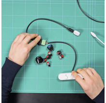 Hände montieren verschiedene Druckschalter und Elektronikbauteile auf einer grünen Schneidematte für ein Bastelprojekt.