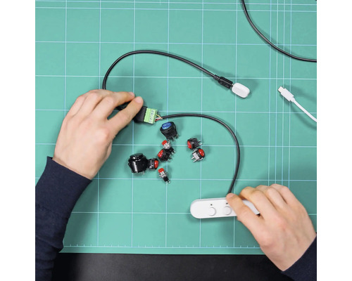 Hände montieren verschiedene Druckschalter und Elektronikbauteile auf einer grünen Schneidematte für ein Bastelprojekt.