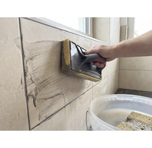 Eine Person reinigt beigefarbene Wandfliesen mit einem Schwammbrett. Ein weißer Eimer mit Wasser steht im Vordergrund.