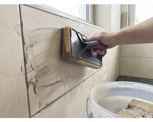 Eine Person reinigt beigefarbene Wandfliesen mit einem Schwammbrett. Ein weißer Eimer mit Wasser steht im Vordergrund.