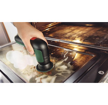 Eine Hand reinigt die Glasscheibe einer Backofentür mit einer elektrischen Akku-Reinigungsbürste und Reinigungsschaum.