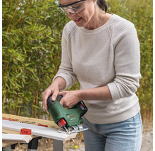 Frau mit Schutzbrille schneidet eine weiße Holzleiste mit einer kabellosen Bosch Stichsäge EasySaw 18 Volt auf einer Werkbank im hellen Garten.