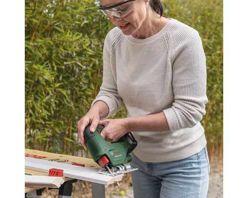 Frau mit Schutzbrille schneidet eine weiße Holzleiste mit einer kabellosen Bosch Stichsäge EasySaw 18 Volt auf einer Werkbank im hellen Garten.