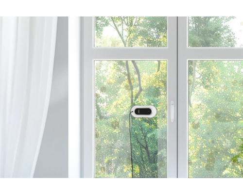 Fensterreinigungsroboter an einer Glasscheibe in einem hellen Raum mit Blick auf Bäume im Freien.
