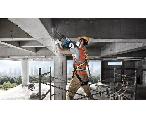 Bauarbeiter mit Schutzausrüstung nutzt Bosch Bohrhammer an einer Betonstruktur auf einer Baustelle mit Gerüst und Stadtansicht.