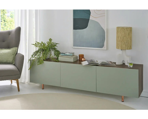 Helles, modern eingerichtetes Wohnzimmer mit einem Sideboard in Grün und Holzoptik, einem grauen Sessel, einer Zimmerpflanze und einer Tischleuchte.
