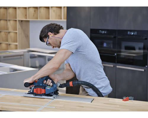 Mann nutzt eine Handkreissäge mit Führungsschiene und Staubabsaugung auf einer Holzoberfläche in einer Küche.