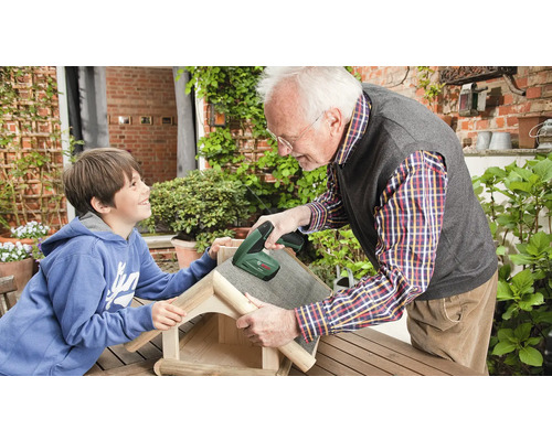 Ein Mann und ein Kind bauen in einem hellen Garten ein Vogelhaus aus Holz mit einem Akku-Tacker von Bosch.