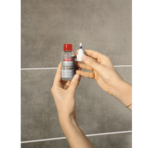 Hände halten eine Flasche Lugato Fugen-Perle Fugenschutz und den dazugehörigen Applikator vor einer gefliesten Wand.