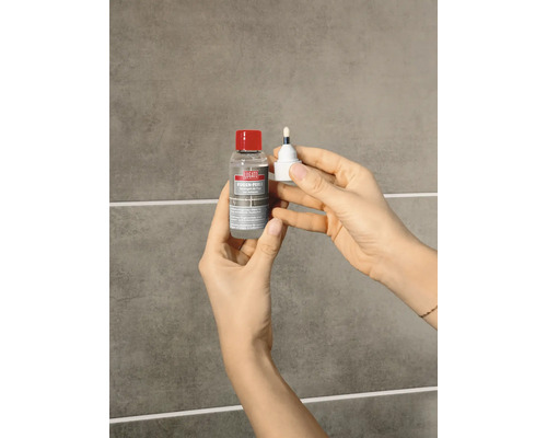 Hände halten eine Flasche Lugato Fugen-Perle Fugenschutz und den dazugehörigen Applikator vor einer gefliesten Wand.
