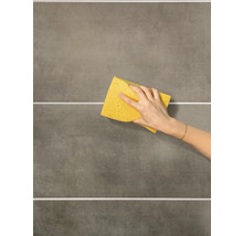 Eine Hand reinigt graue Wandfliesen mit einem gelben Schwamm.