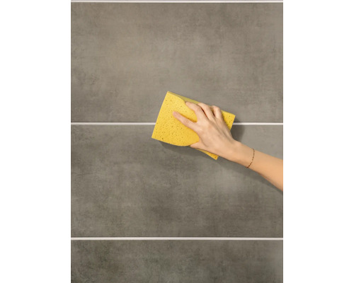 Eine Hand reinigt graue Wandfliesen mit einem gelben Schwamm.