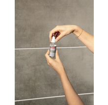 Hände halten eine Flasche Lugato Fugen-Perle vor einer gefliesten Wand zur Anwendung auf Fliesenfugen.