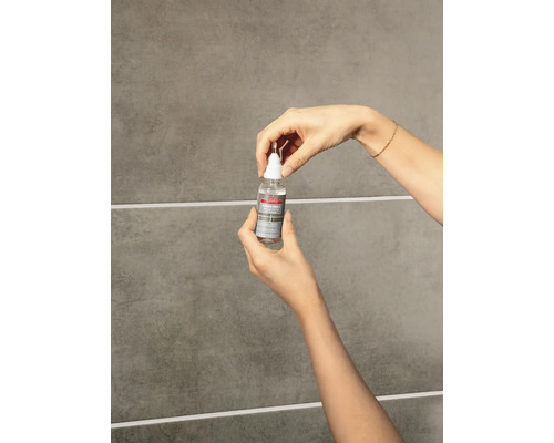 Hände halten eine Flasche Lugato Fugen-Perle vor einer gefliesten Wand zur Anwendung auf Fliesenfugen.