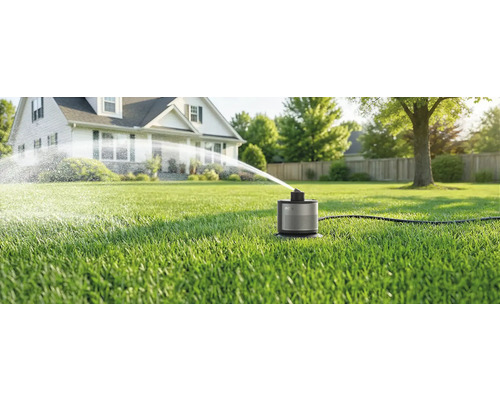 Kompakter smarter Rasensprenger auf grünem Rasen sprüht Wasserbogen vor einem Wohnhaus im hellen Garten.