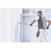 Handwerker in Arbeitskleidung streicht eine Wand mit einer Farbwalze und einer Teleskopstange in einem hellen Innenraum.