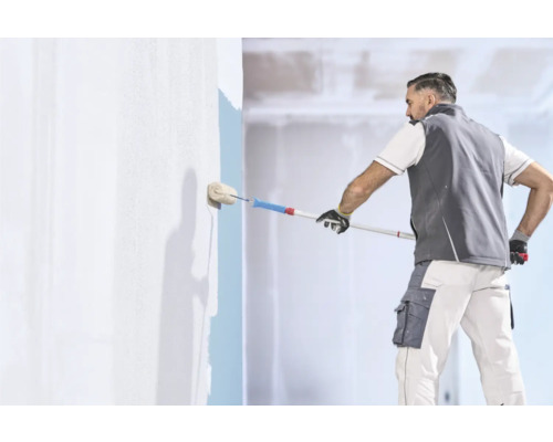 Handwerker in Arbeitskleidung streicht eine Wand mit einer Farbwalze und einer Teleskopstange in einem hellen Innenraum.