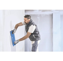 Handwerker glättet eine weiße Wand mit einem breiten blauen Flächenspachtel in einem hellen Raum.