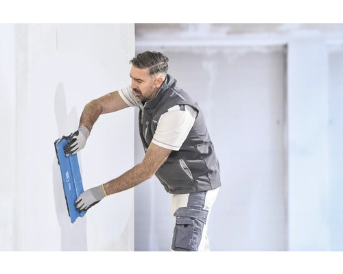 Handwerker glättet eine weiße Wand mit einem breiten blauen Flächenspachtel in einem hellen Raum.