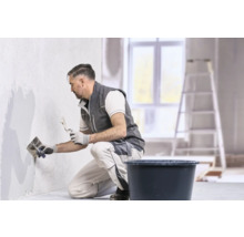 Ein Handwerker in Arbeitskleidung trägt mit einer Glättkelle und einem Spachtel Putz auf eine Wand auf.