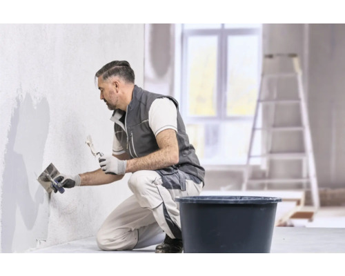 Ein Handwerker in Arbeitskleidung trägt mit einer Glättkelle und einem Spachtel Putz auf eine Wand auf.