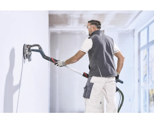 Handwerker glättet eine Wand mit einem Langhalsschleifer in einem hellen Innenraum.