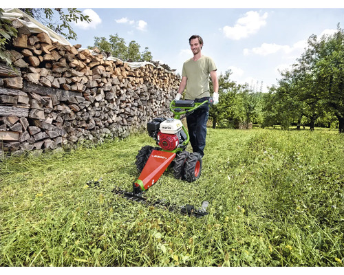 Ein Mann bedient einen agria Balkenmäher auf einer hellen Wiese mit hohem Gras vor einem Holzstapel in einem Garten.