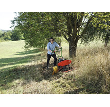 Ein Mann bedient einen Balkenmäher auf einer Wiese mit hohem Gras unter einem Baum.