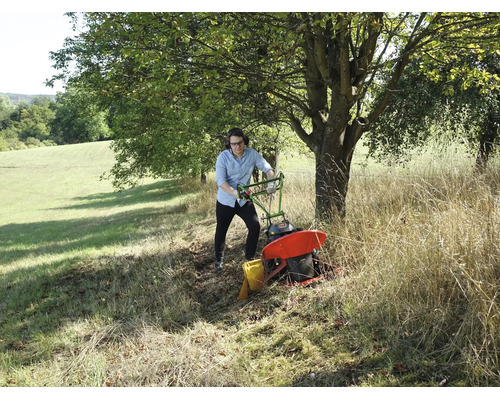 Ein Mann bedient einen Balkenmäher auf einer Wiese mit hohem Gras unter einem Baum.