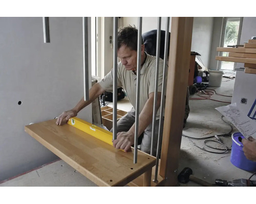 Handwerker beim Ausrichten einer hölzernen Treppenstufe mit einer Wasserwaage an einem Metallgeländer in einem hellen, im Bau befindlichen Raum.