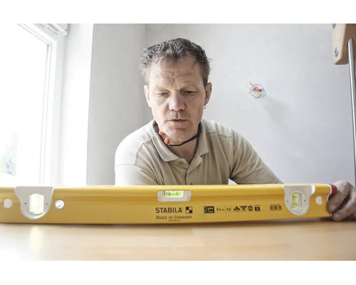 Handwerker betrachtet eine gelbe Stabila Wasserwaage Typ R 300 mit einer Länge von 61 Zentimetern auf einem Tisch in einem hellen Raum.