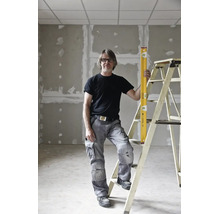 Handwerker in grauer Arbeitshose und schwarzem T-Shirt steht mit einer gelben Wasserwaage an einer Leiter im Rohbau.