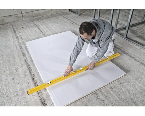 Handwerker schneidet eine Gipskartonplatte auf dem Betonboden mit einer Wasserwaage und einem Cuttermesser im Innenausbau zu.