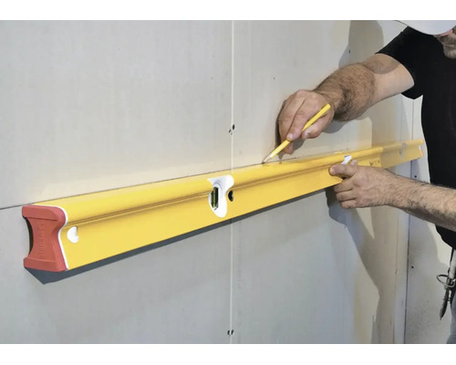 Eine Person hält eine lange Wasserwaage an eine Trockenbauwand und markiert mit einem Bleistift eine waagerechte Linie.