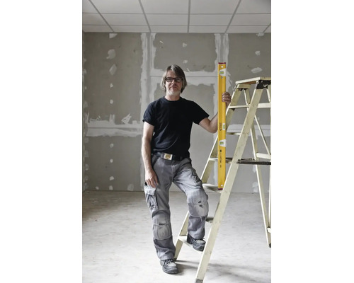Handwerker in grauer Arbeitshose und schwarzem T-Shirt steht mit einer gelben Wasserwaage an einer Leiter im Rohbau.