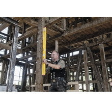 Ein Handwerker in Zunftkleidung prüft mit einer langen gelben Wasserwaage die Ausrichtung eines Holzbalkens in einem Fachwerkbau.