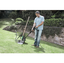 Ein Mann reinigt eine verschmutzte Schaufel in einer Schubkarre mit einem Hochdruckreiniger auf einer Rasenfläche im Garten.