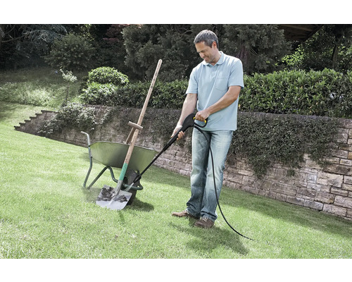 Ein Mann reinigt eine verschmutzte Schaufel in einer Schubkarre mit einem Hochdruckreiniger auf einer Rasenfläche im Garten.