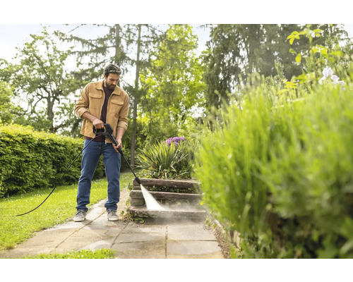 Ein Mann reinigt Steinstufen im Garten mit einem Hochdruckreiniger in einer hellen Umgebung.
