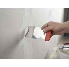 Eine Hand trägt mit einem Malerspachtel aus Edelstahl Spachtelmasse auf eine Wandfuge auf.