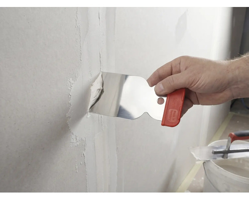 Eine Hand trägt mit einem Malerspachtel aus Edelstahl Spachtelmasse auf eine Wandfuge auf.