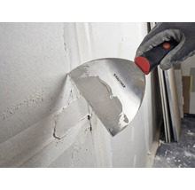 Hand in einem Arbeitshandschuh trägt mit einer Rautner Malerspachtel Spachtelmasse auf eine Wand auf.