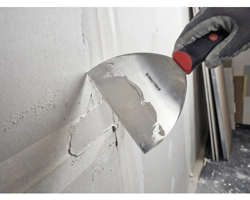Hand in einem Arbeitshandschuh trägt mit einer Rautner Malerspachtel Spachtelmasse auf eine Wand auf.