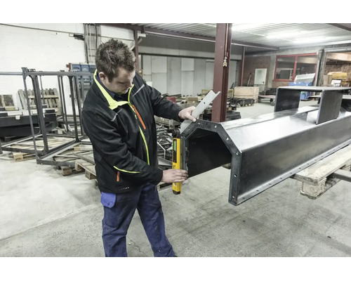 Handwerker in einer Werkstatt verwendet einen digitalen Winkelmesser, um eine große Metallkonstruktion auf einer Palette präzise zu messen.