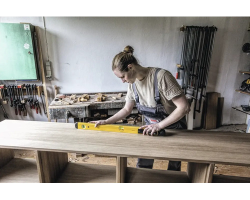 Handwerkerin prüft mit einer Wasserwaage die Ausrichtung einer Holzplatte in einer gut organisierten Werkstatt.