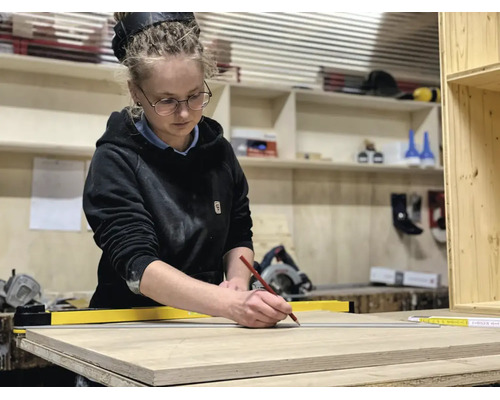 Eine Frau in einer Werkstatt markiert eine Holzplatte mit einem Bleistift und einer gelben Richtlatte. Im Hintergrund sind Regale mit Werkzeugen.