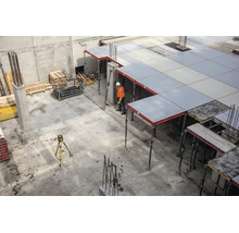 Baustelle mit Deckenschalung aus grauen Paneelen, Baustützen, einem Bauarbeiter in Warnkleidung und einem Nivelliergerät auf einem Stativ.